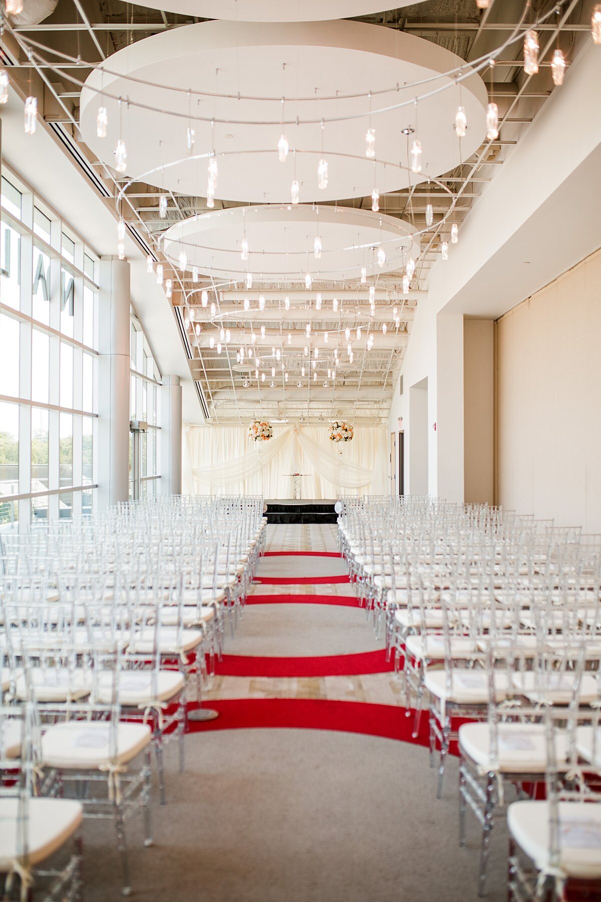 Wedding ceremony at Liff Center with clear chivari chairs with white cushions. The red and white carpet lead up to a white and orange ceremony space with crystal chandeliers at the Nashville Opera - Noah Liff Opera Center