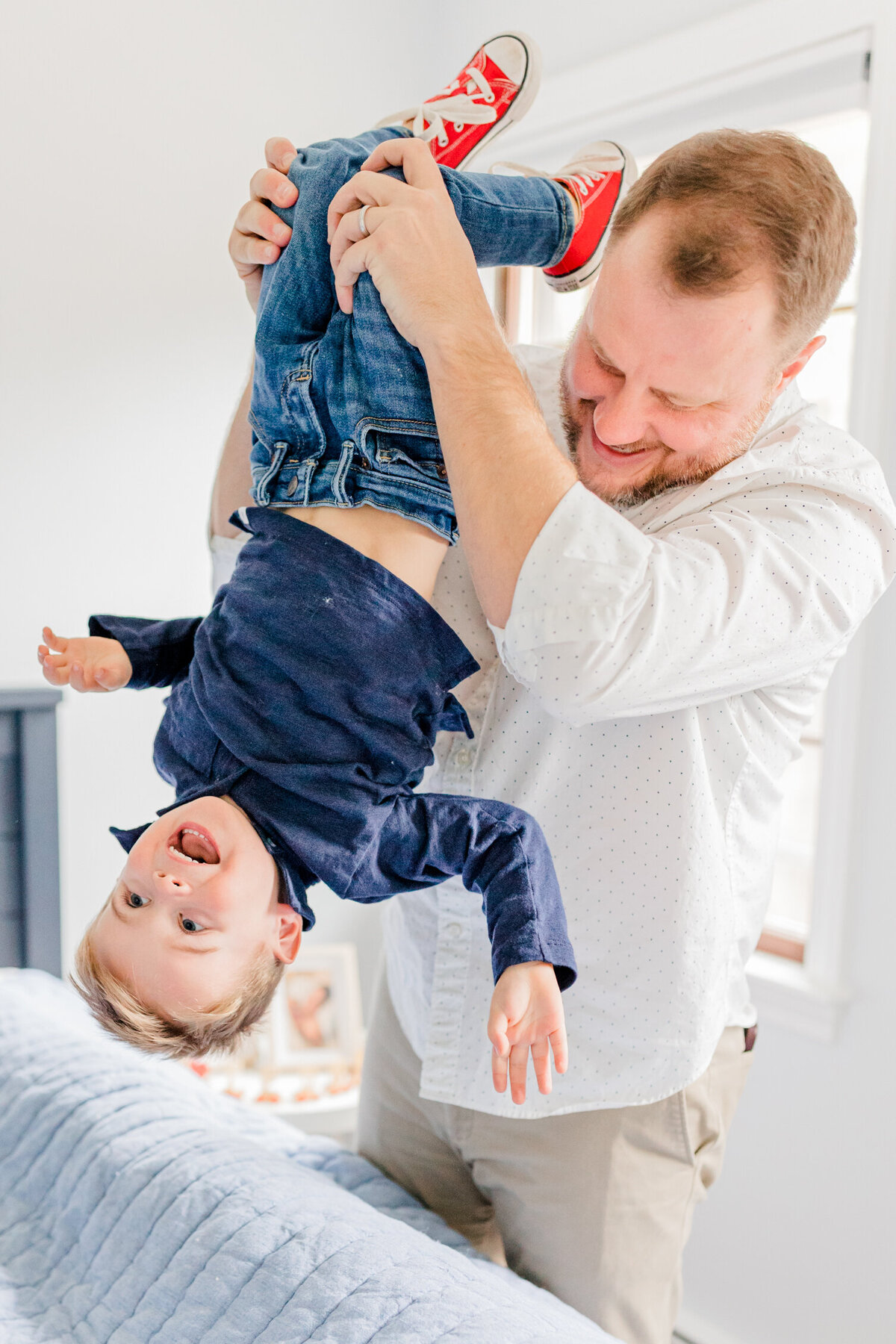 Dad in white shirt holds laughing toddler in blue shirt upside-down
