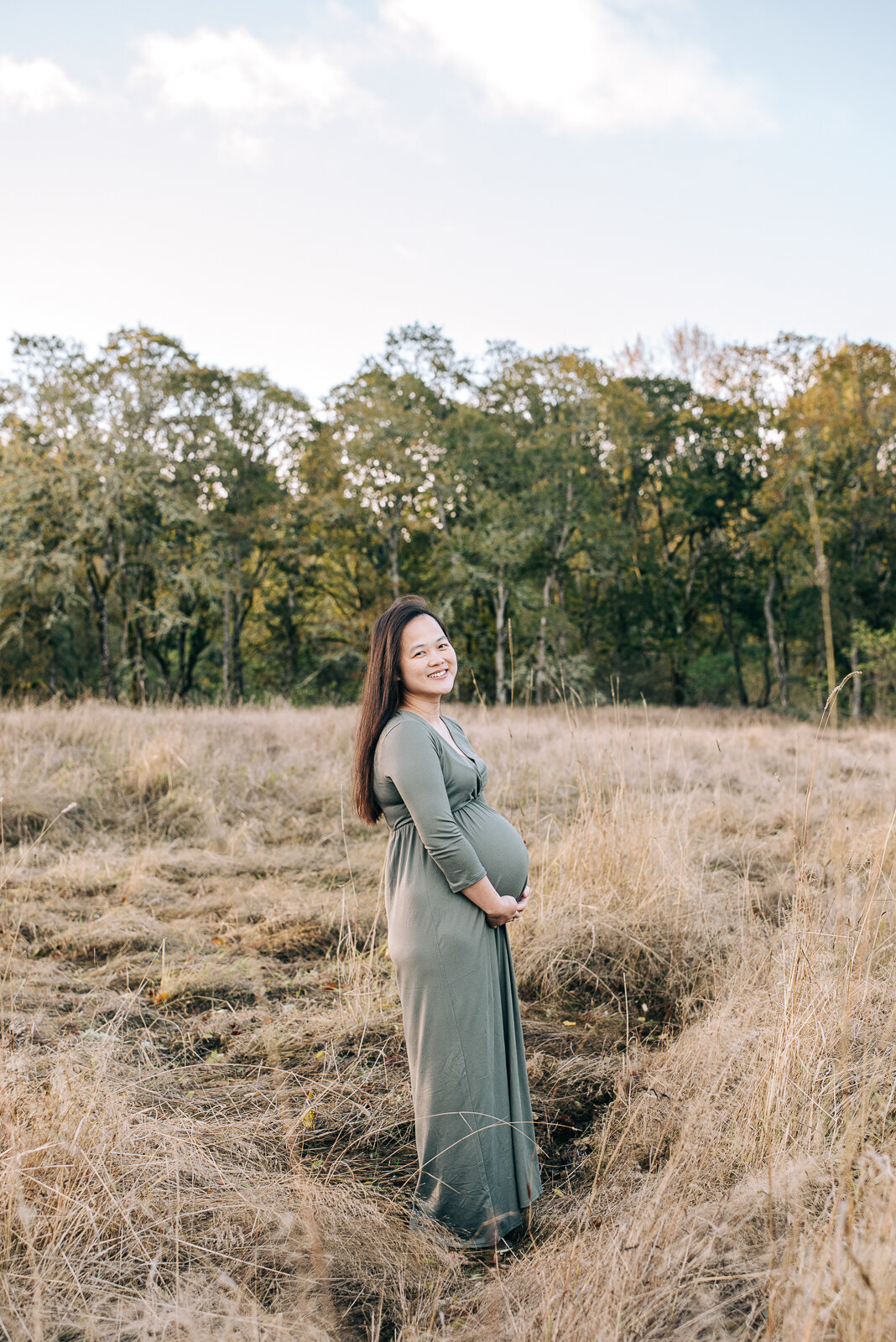 Pregnant mom wearing a sage green long sleeve maternity dress smiling at the camera in a field at Fort Steilacoom Park