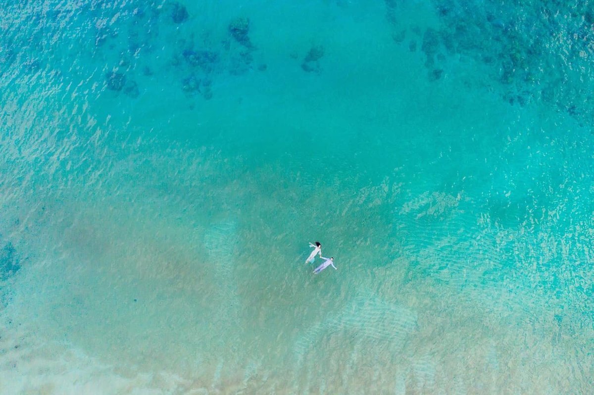Couple hold hands and are photographed with a drone during their beach engagement session with Love + Water