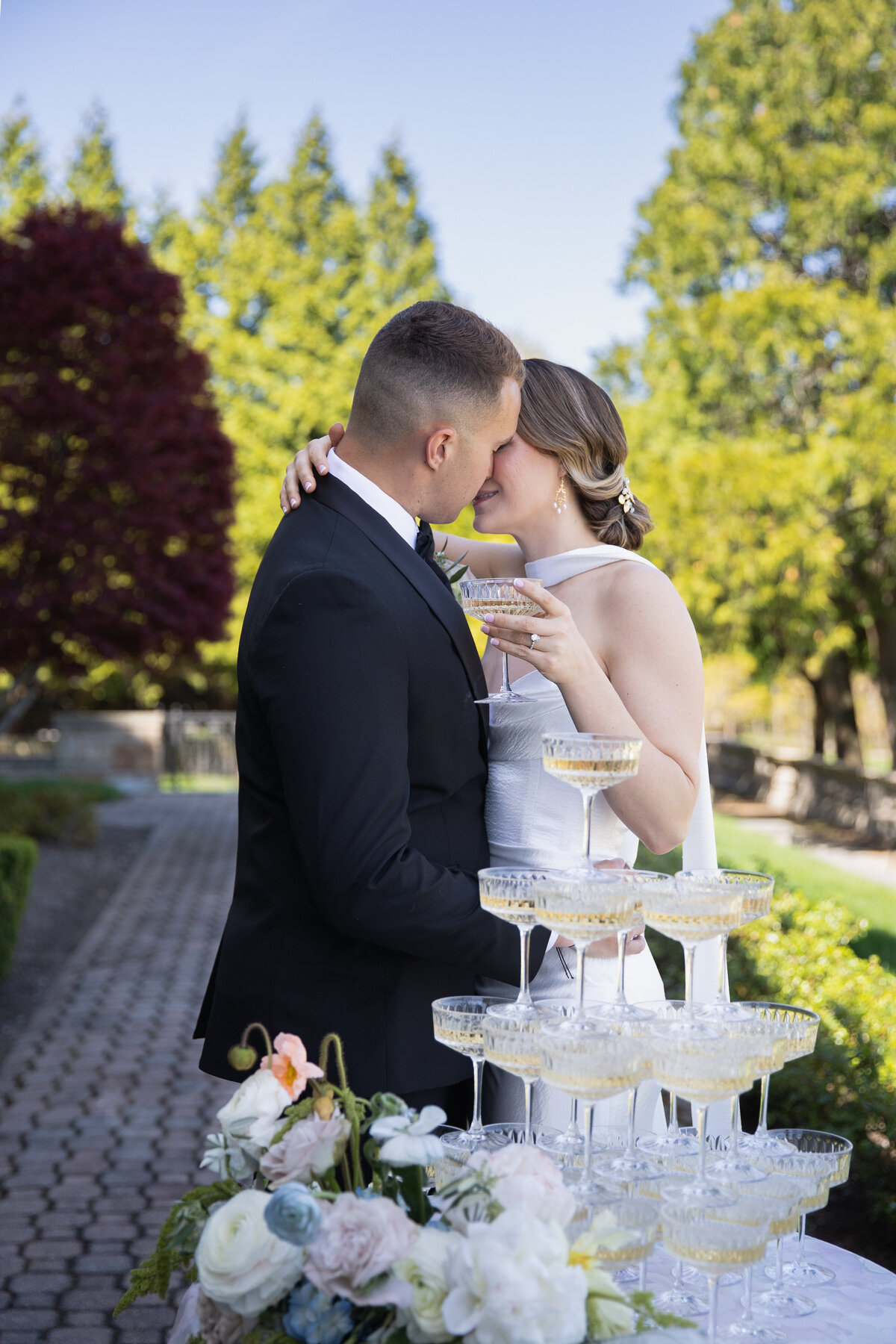 Rhode Island Wedding Photographers | Kelsey Sheehan Photography Timeless Rhode Island Weddings | A couple lovingly embraces in a sunlit garden, holding champagne glasses beside a stacked pyramid. A serene, joyful atmosphere with lush greenery surrounds them.