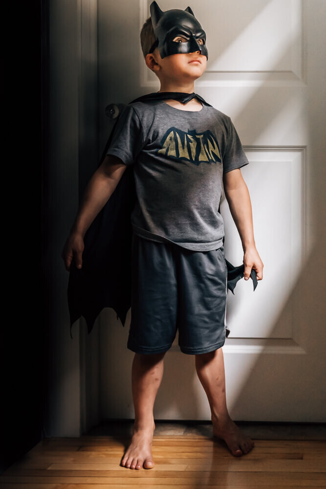 A small boy dressed in a batman mask and cape, gazing outa window toward dramatic lighting.