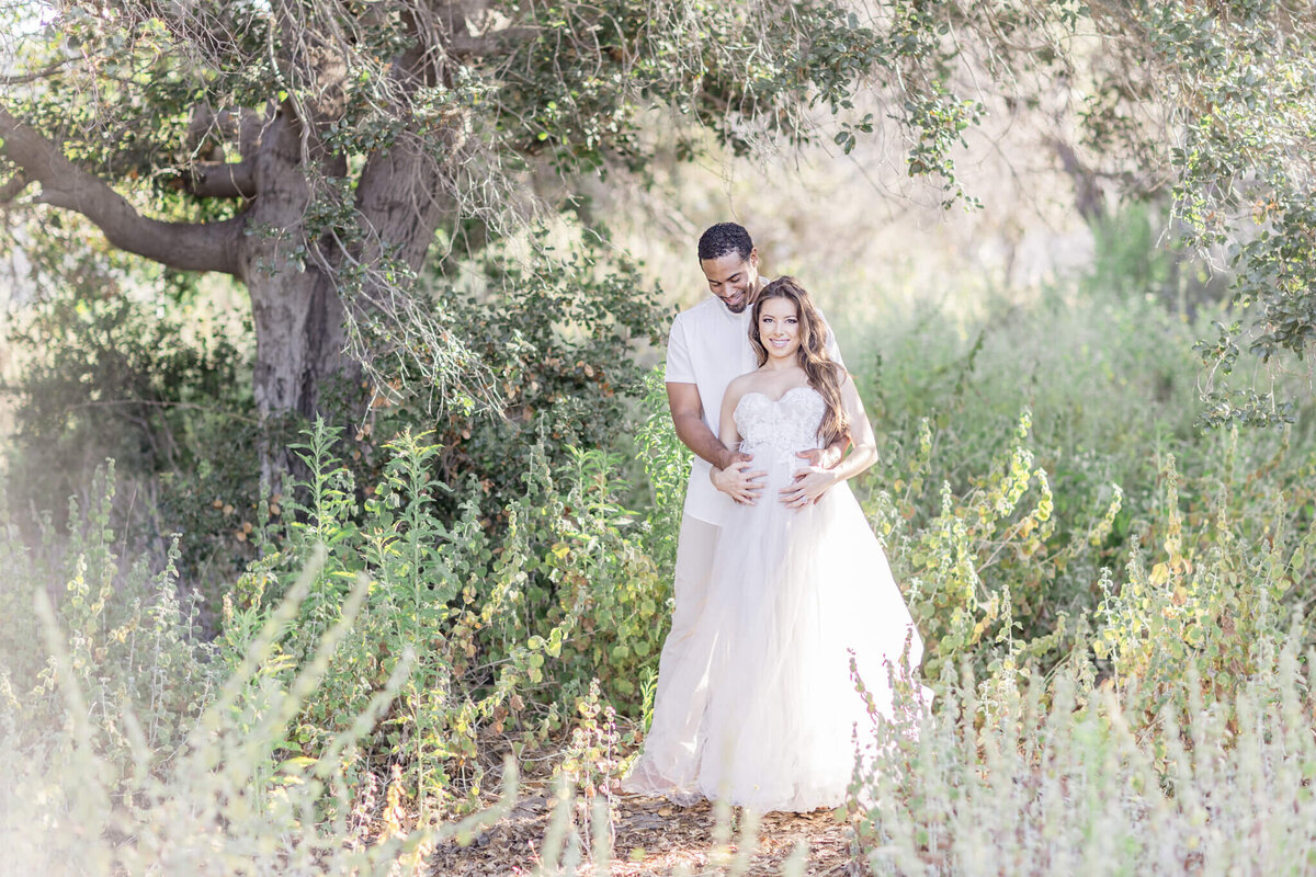 A couple stands in a sunlit forest clearing. The woman, in a flowing white dress, is embraced from behind by the man, who is wearing a white shirt. They are surrounded by tall green plants and trees, creating a serene and romantic atmosphere.