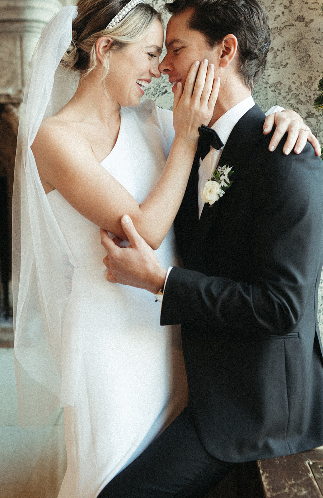 A bride and groom smile and hold each other closely, with the bride touching the groom's face. The bride is in a white dress and veil, and the groom is in a black suit with a white boutonnière. Their love shines brightly against the stunning backdrop of their Banff wedding.