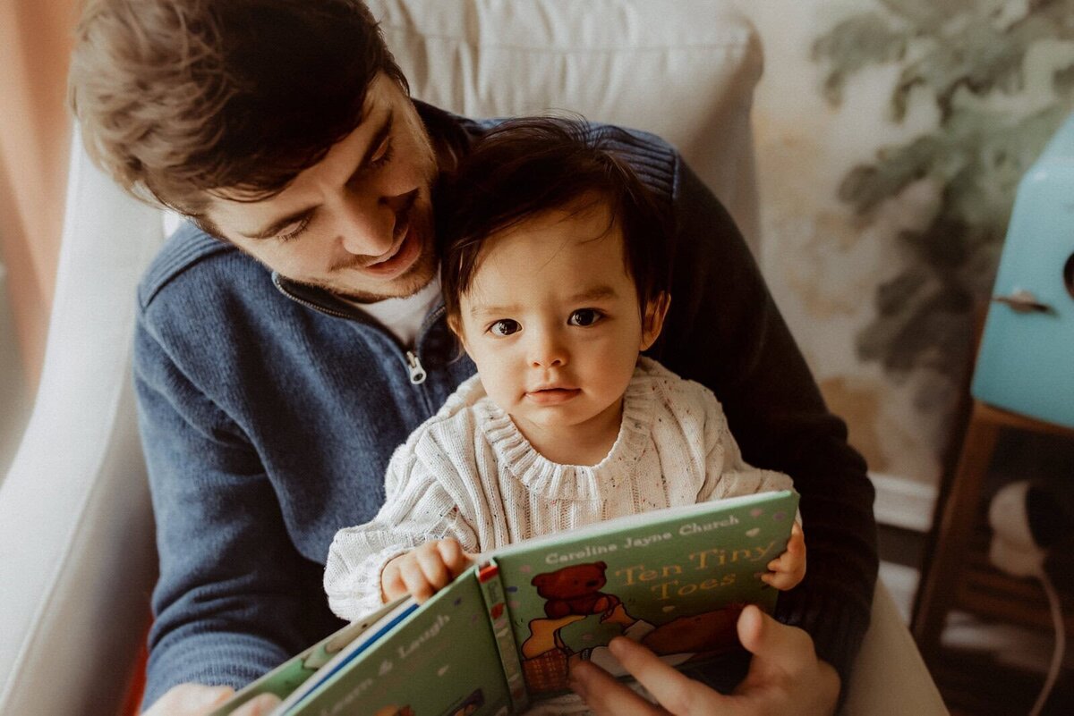 Man and child sitting in a chair, reading "Ten Tiny Toes" together. The child is wearing a pale sweater and the man is in a dark zippered top. A cherished moment captured by a family photographer in Warner Robins, GA.