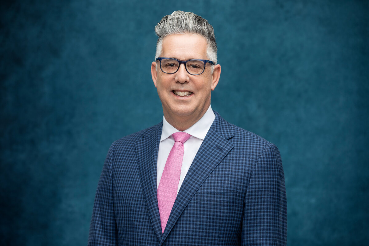 A Caucasian white man with silver white hair wearing a blue check jacket and a pink tie smiles for a professional headshot photo at Janel Lee Photography studios in Cincinnati Ohio