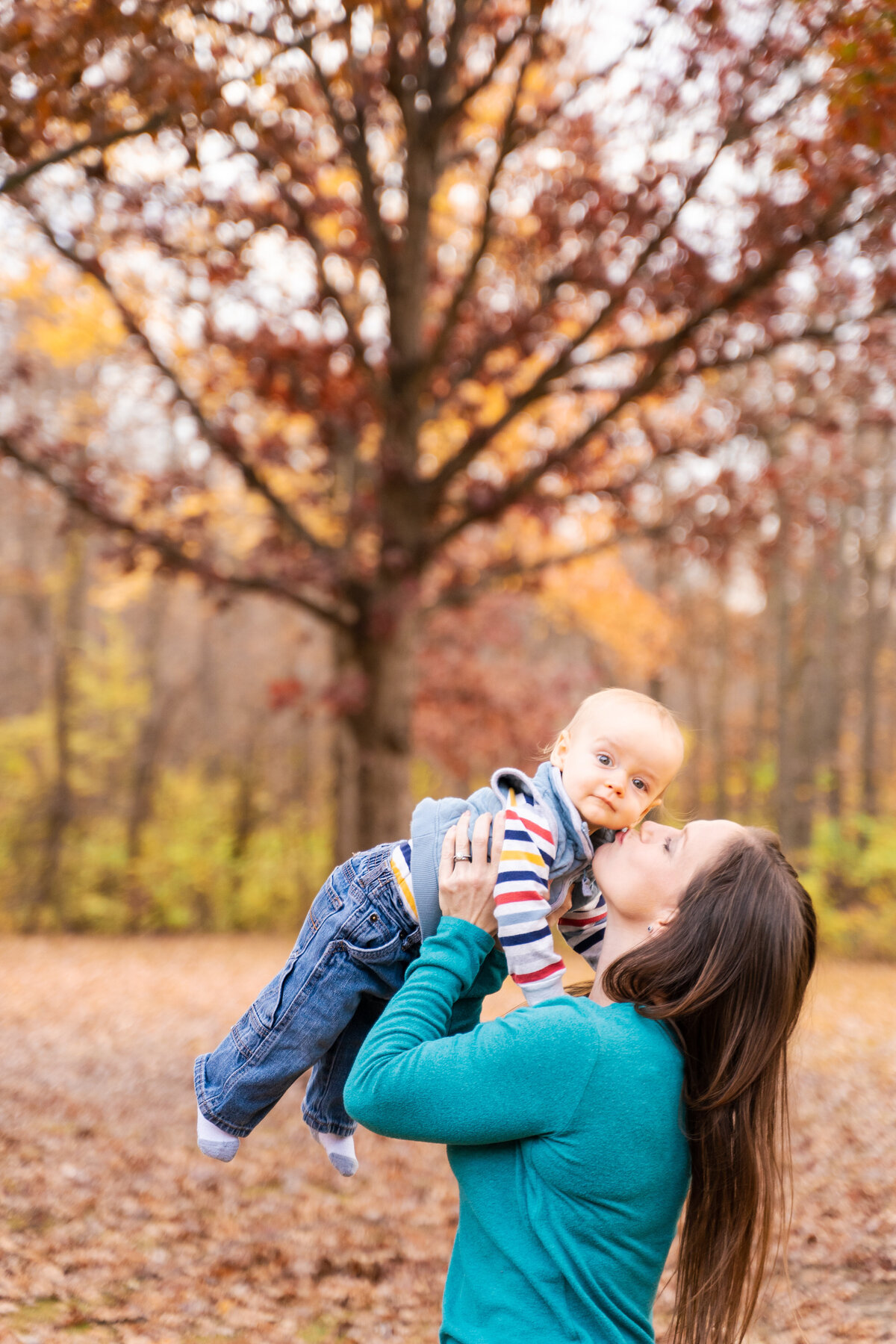 Mother holds up her baby and kisses him on the check in front of a colorful tree - Sharon Woods Westerville, Ohio