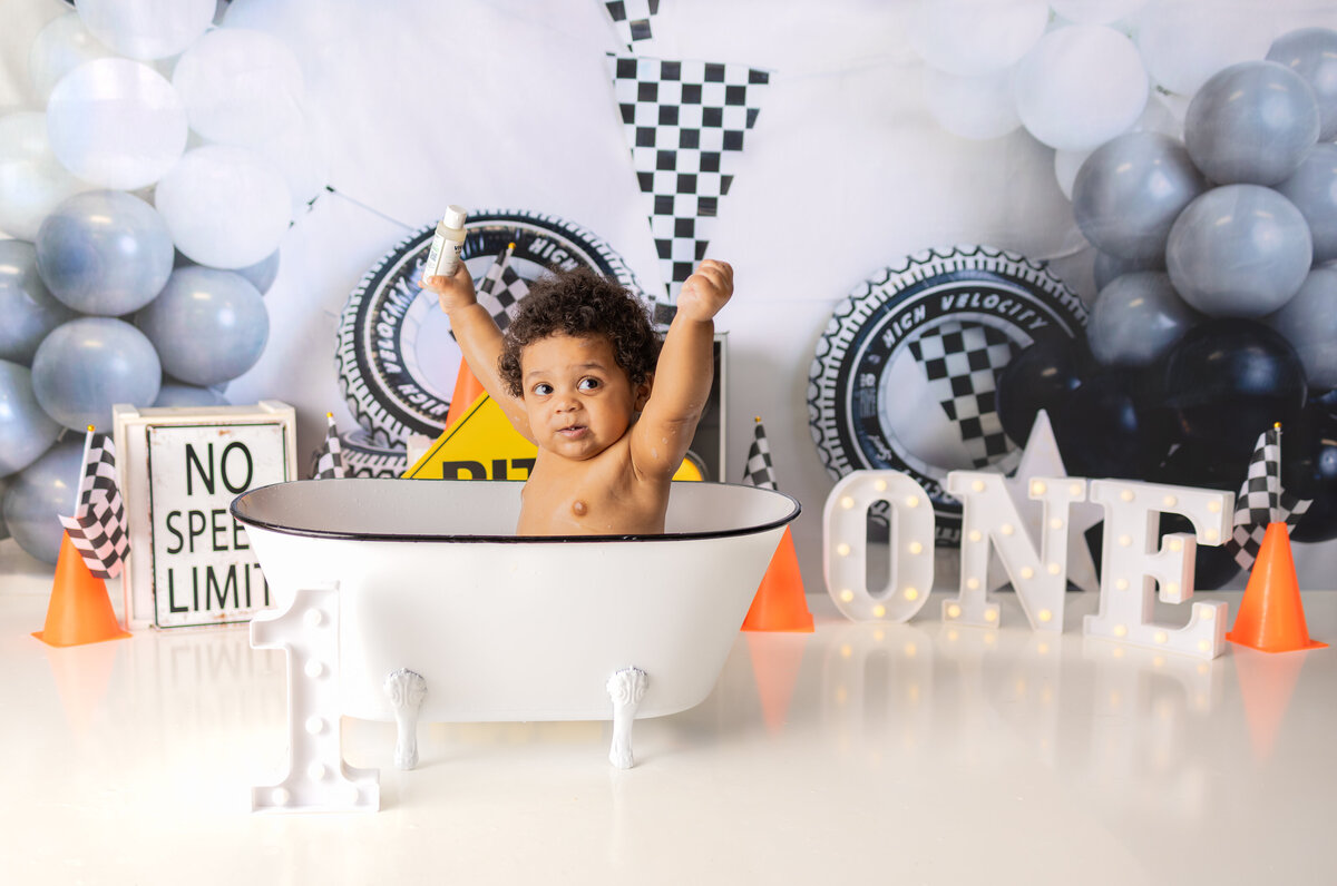 Happy baby boy celebrating his first birthday in race car themed bath setup with arms raised after cake smash in Waldwick NJ studio