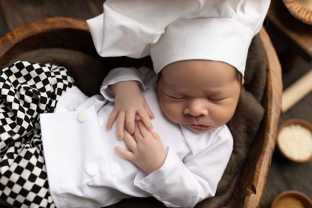 Close-up of a sleeping newborn in a white chef hat and coat, nestled in a heart-shaped wooden bowl.