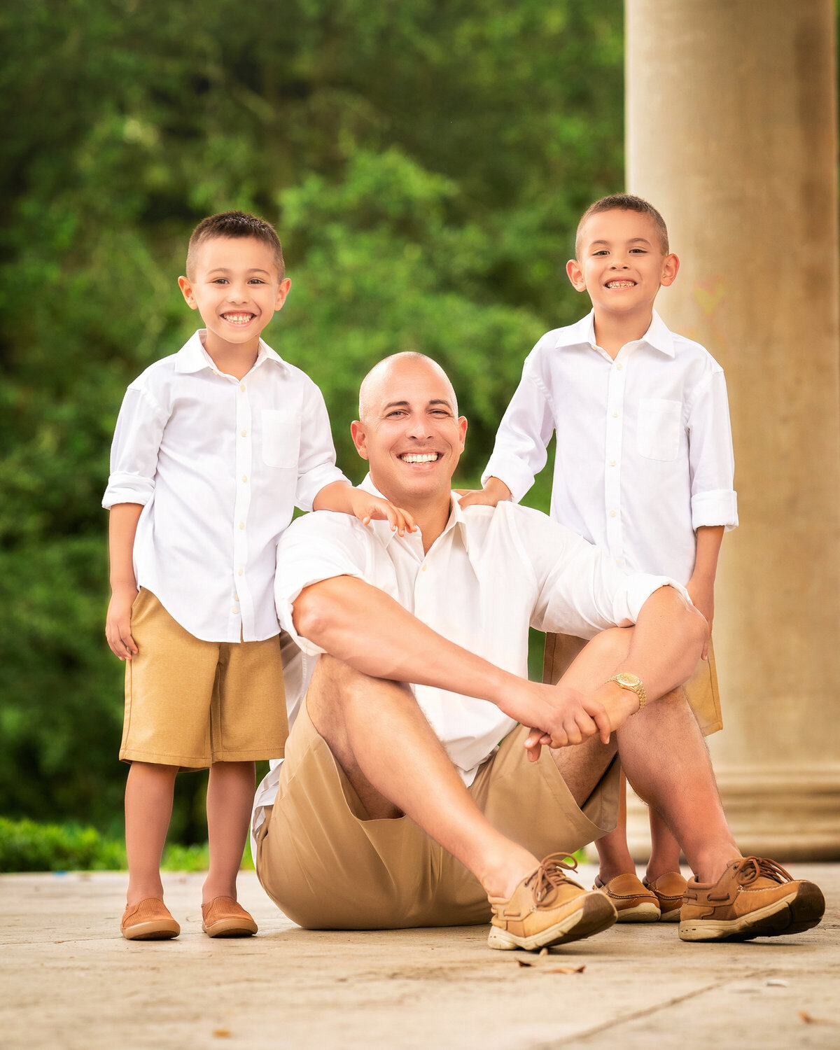 Dad is sitting on the ground with each of his sons standing next to him.  They are wearing white shirts and khaki shorts.  Dad is bald.  They are all smiling at the camera.