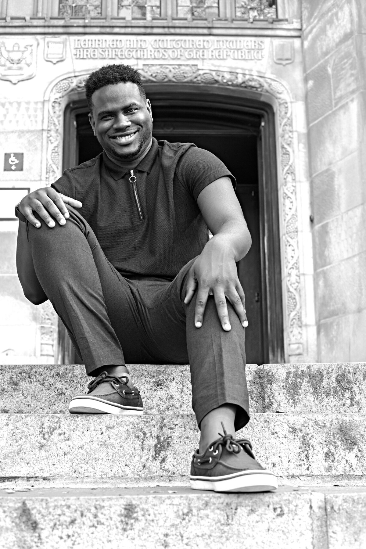 African male wearing a navy blue zip up top, blue pants, and blue Sperry boat shoes sitting on steps in front of library