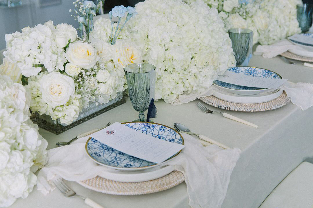a table topped with lots white flowers, luxury wedding menus, and blue and white  dinnerware