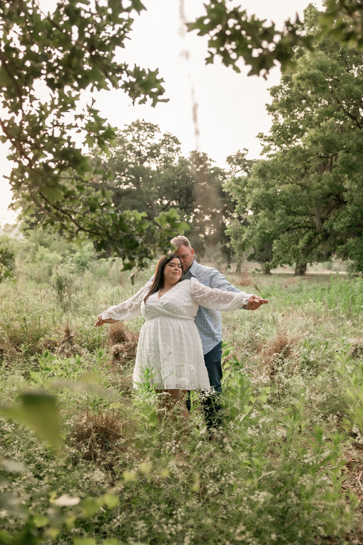 bride and groom sway in a field of grass, while she wears a white engagement dress and he snuggles her.