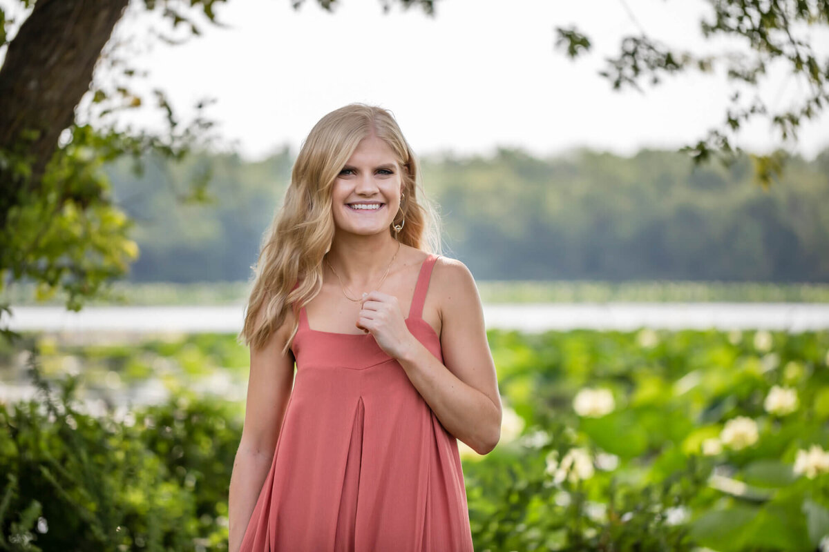 A lovely landscape senior portrait of a beautiful blonde girl wearing a coral dress standing in front of a lake covered in waterlilies. Captured by Springfield, MO senior photographer Dynae Levingston.