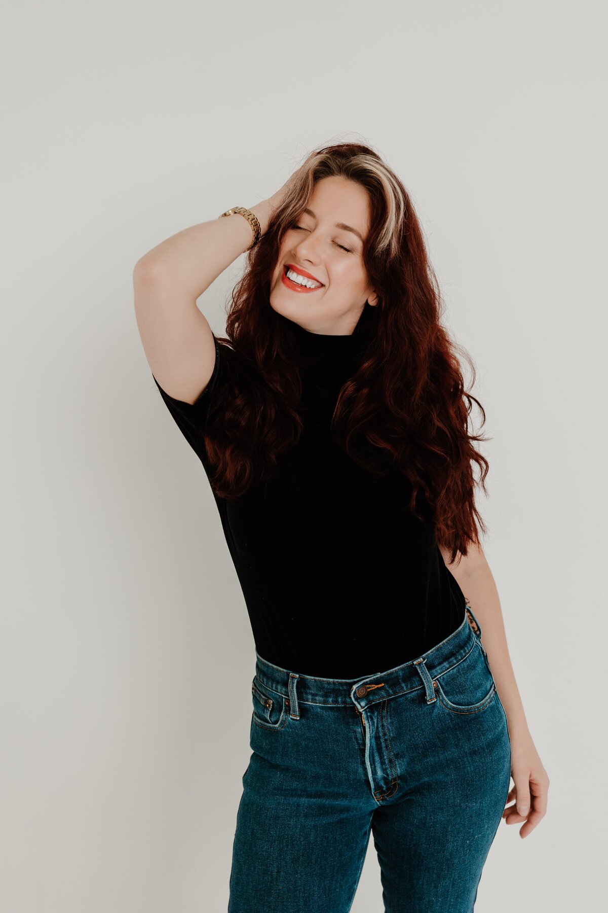 A joyful woman with long wavy red hair, wearing a black turtleneck and blue jeans, playfully posing with her hand in her hair against a plain white background.