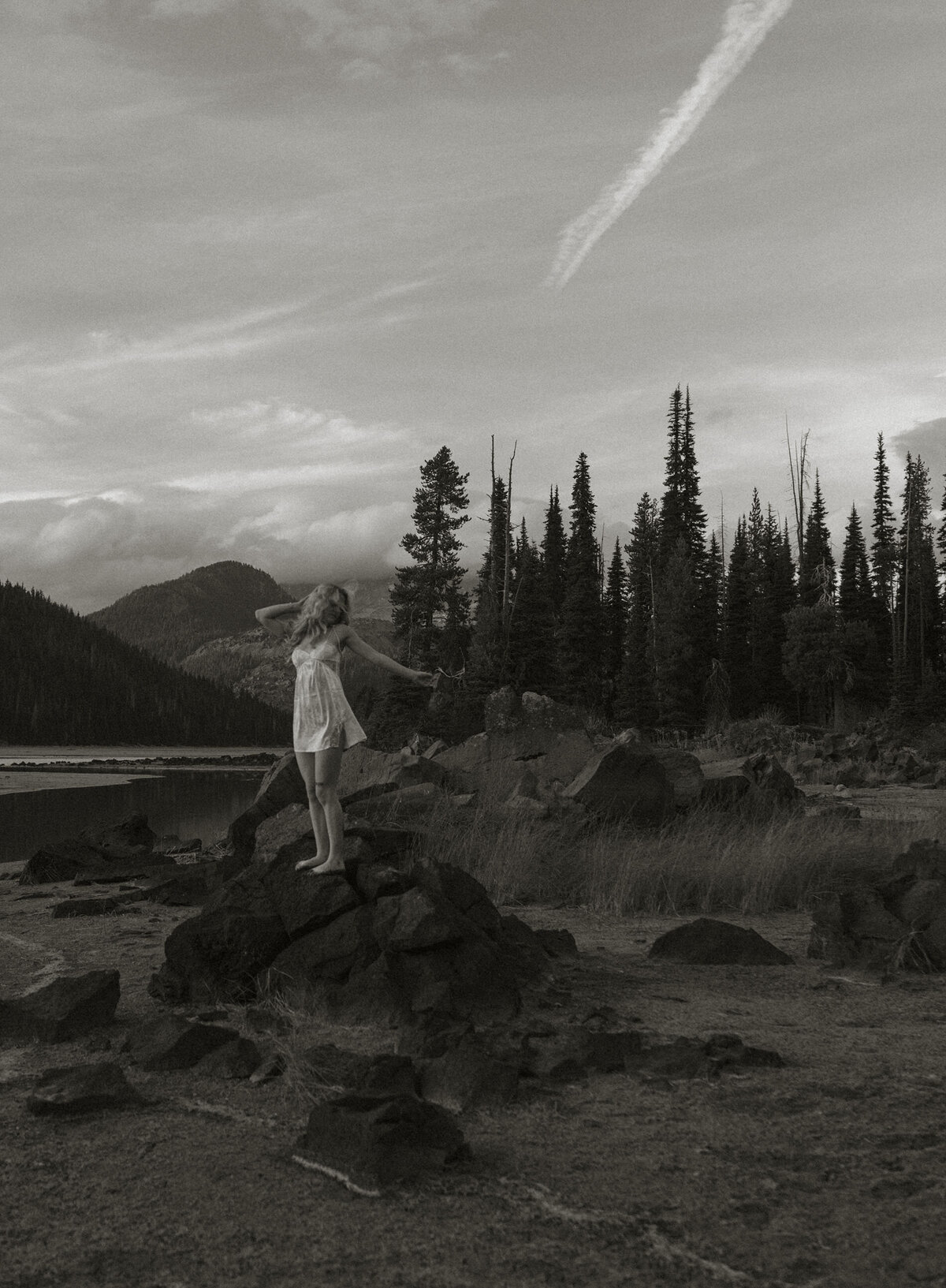 woman in a white dress stands on rock near the lake and looks off into the distance in Oregon