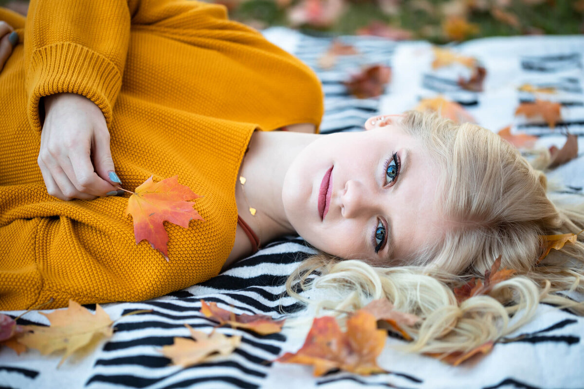 A lovely blonde haired blue eyed senior girl poses laying down on a black and white blanket and surrounded by fallen Fall leaves. Captured by Springfield, MO senior photographer Dynae Levingston.