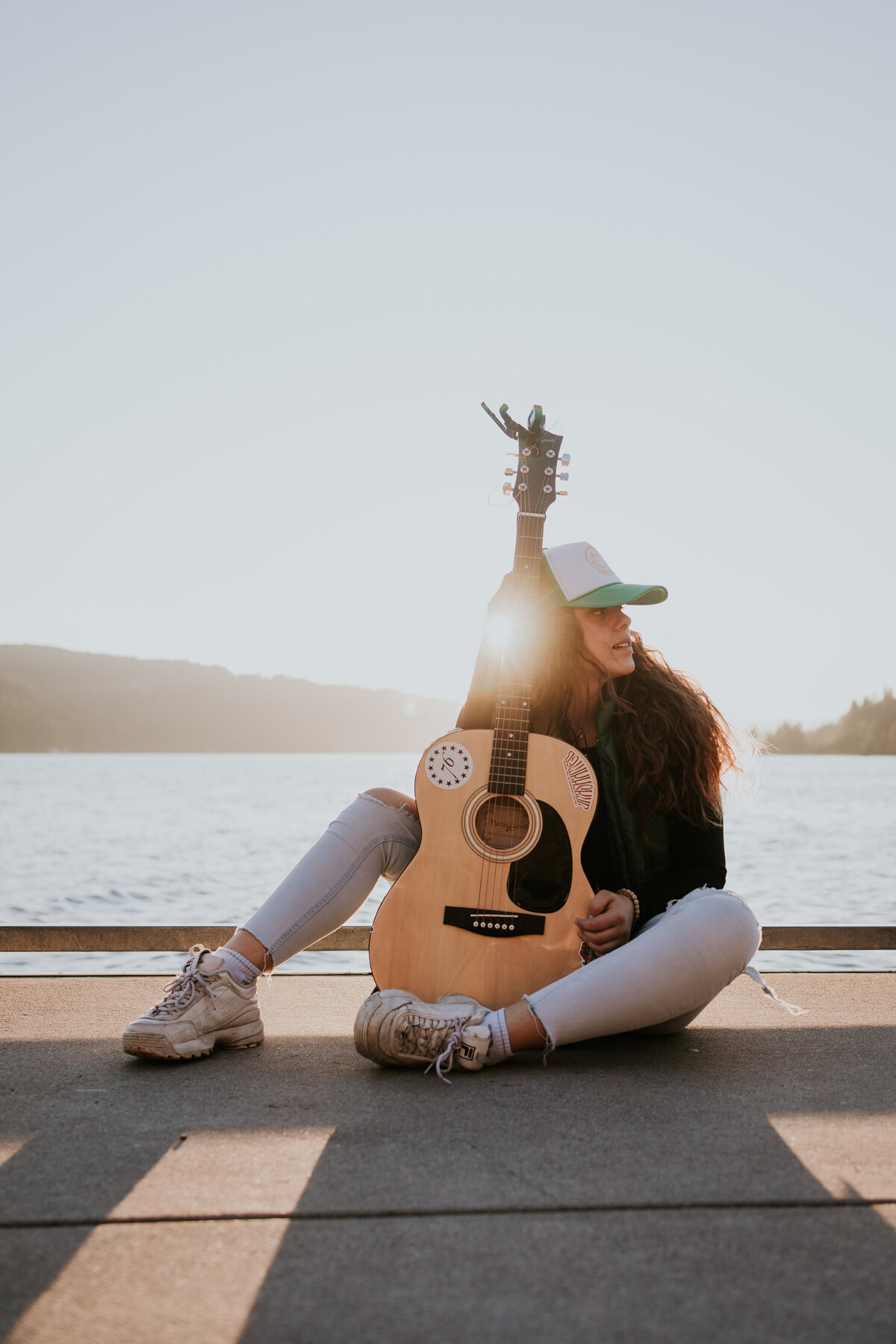 Young woman sits at the edge of dock while holding guitar as sun sets.