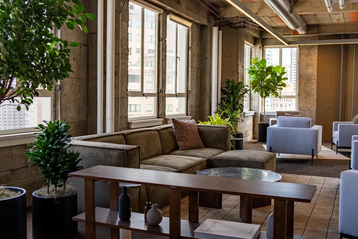 Industrial-style lounge with a large sectional sofa, potted plants, and city views through tall concrete-framed windows.