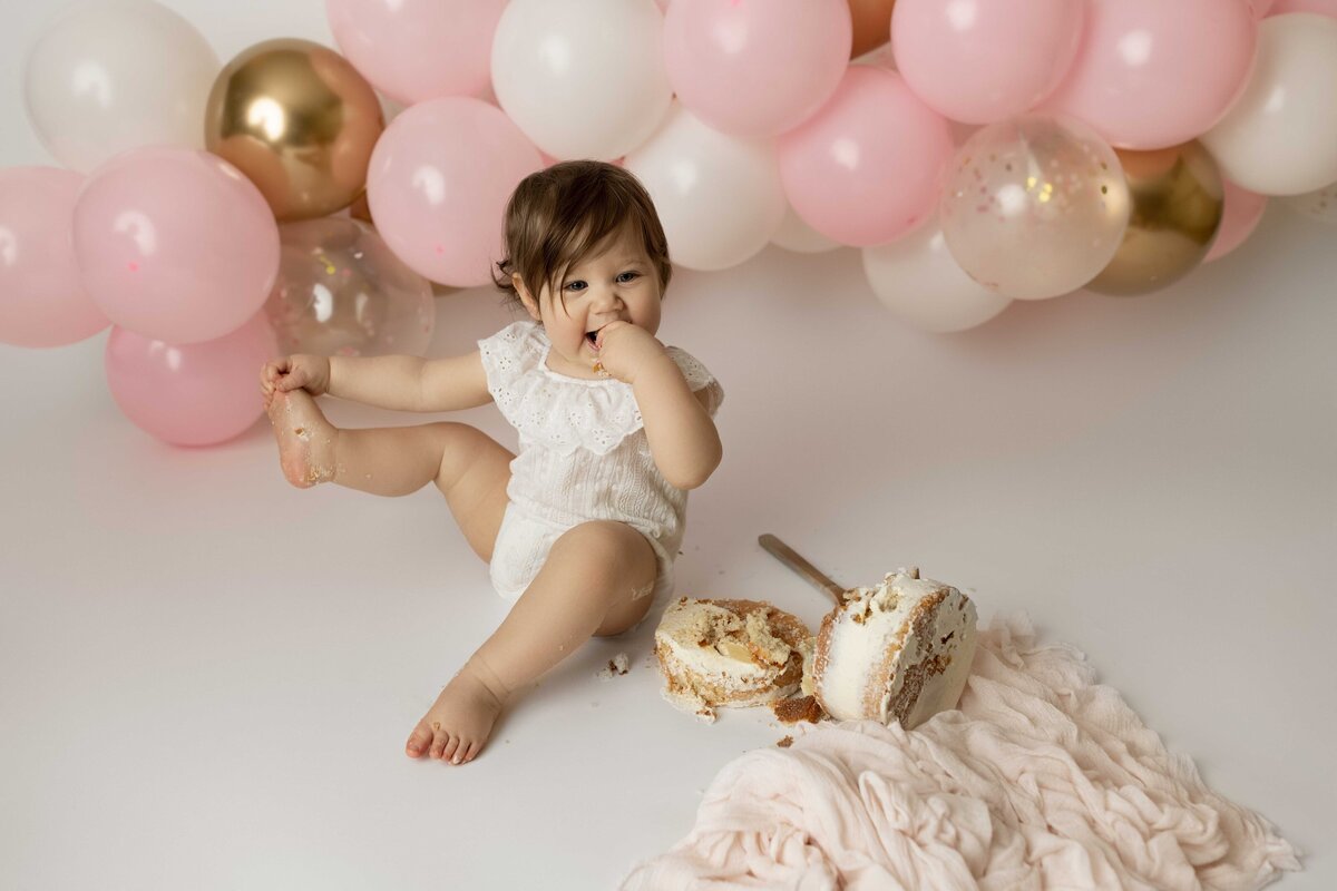 Cake smash photoshoot in London, ON - baby is sitting holding toes with messy cake in front of her. Pink, gold, and cream balloons arch in the background.