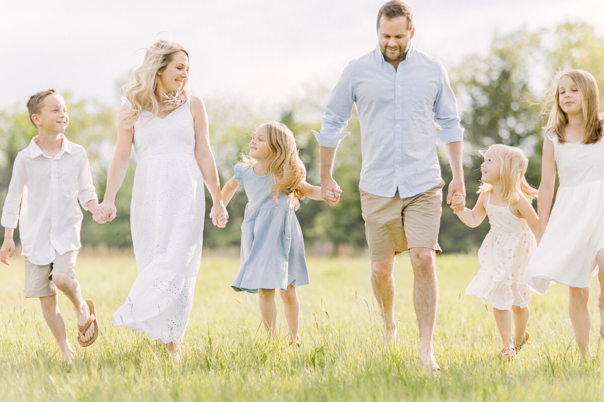 Family of six holding hands and walking across a grassy field for their family photos in Kansas City with photographer Jeanna Hayes