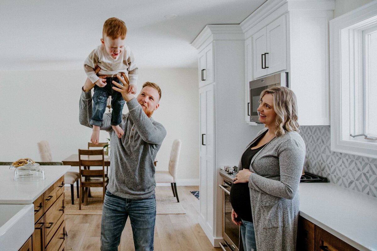 In home maternity session. Expectant mom, and toddler in kitchen. Dad is lifting little boy above his head, mom is smiling at toddler and holding her bump.