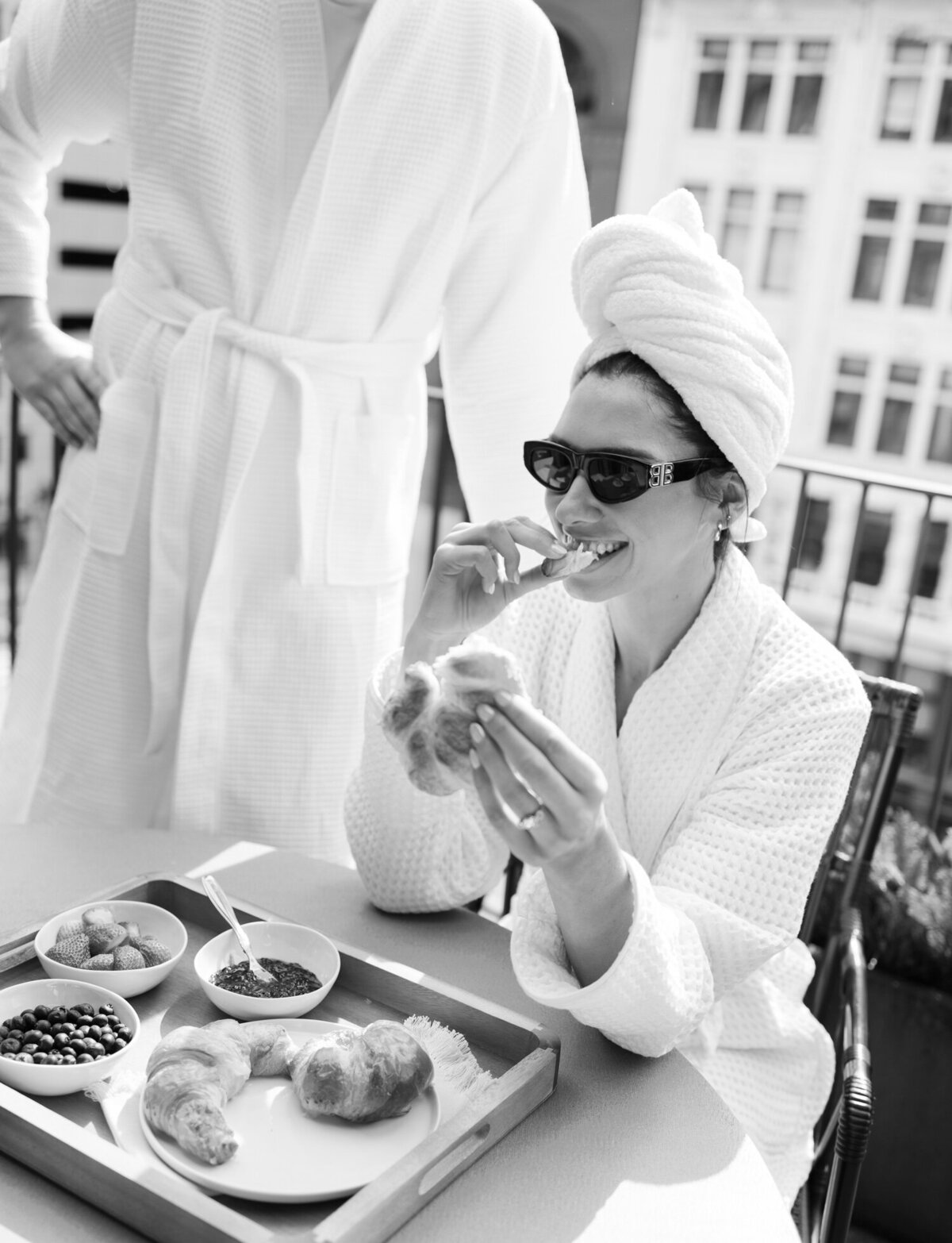 woman laughs eating croissant on Shinola Hotel balcony, couple in bathrobes, sunglasses