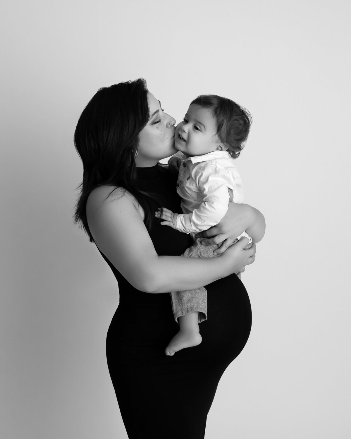 Maternity photo of a mother kissing her toddler while holding him, taken at a Leander, TX photography studio.