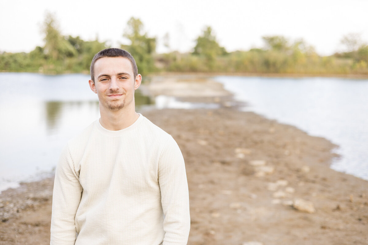 A high school senior male, standing next to a pond, soft smile at the camera, with short brown hair, wearing a light tan long sleeve shirt.