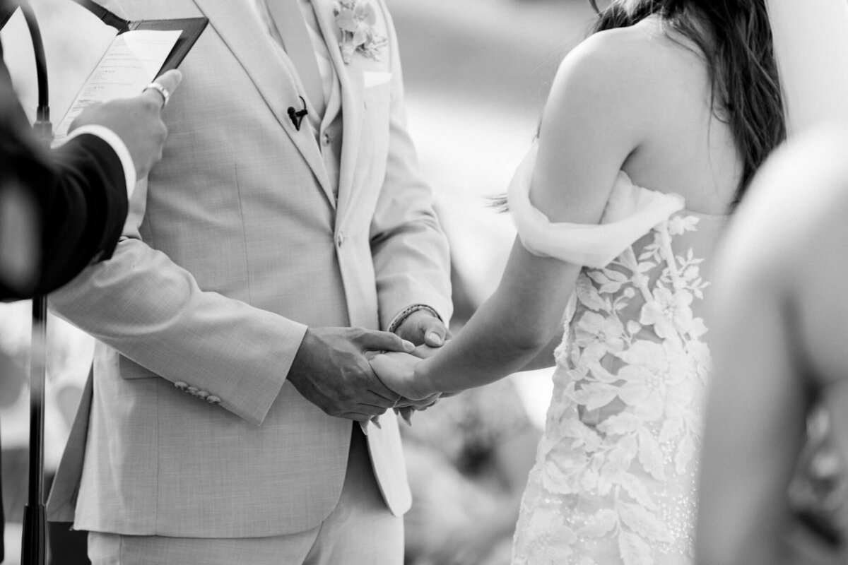 A couple holds hands during a wedding ceremony, with the groom in a light suit and boutonniere and the bride in a lacy dress. A sense of love and commitment.