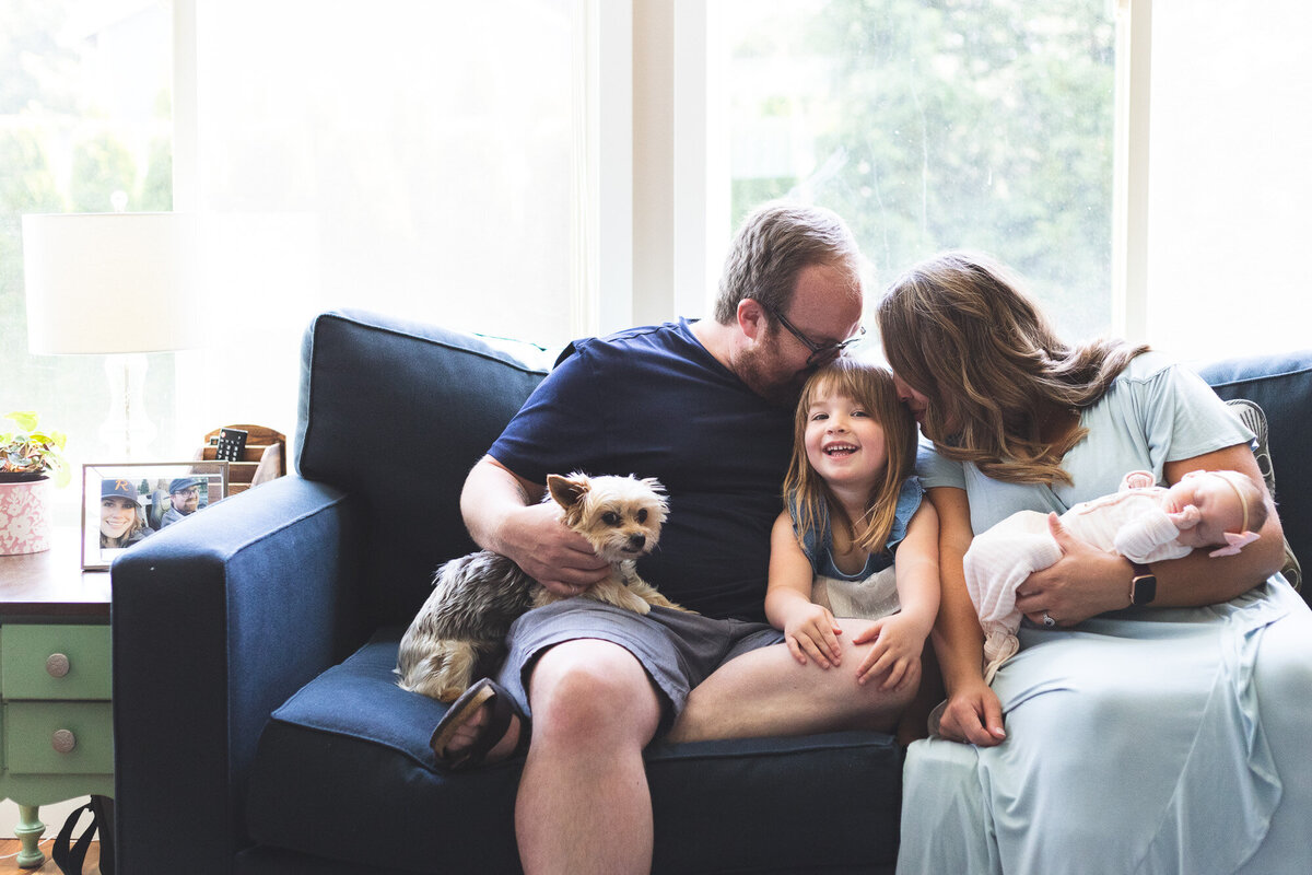 Family of 4 with dog sitting on a blue couch  kissing daughter and newborn baby. Photo by newborn photographer Portland, Or.