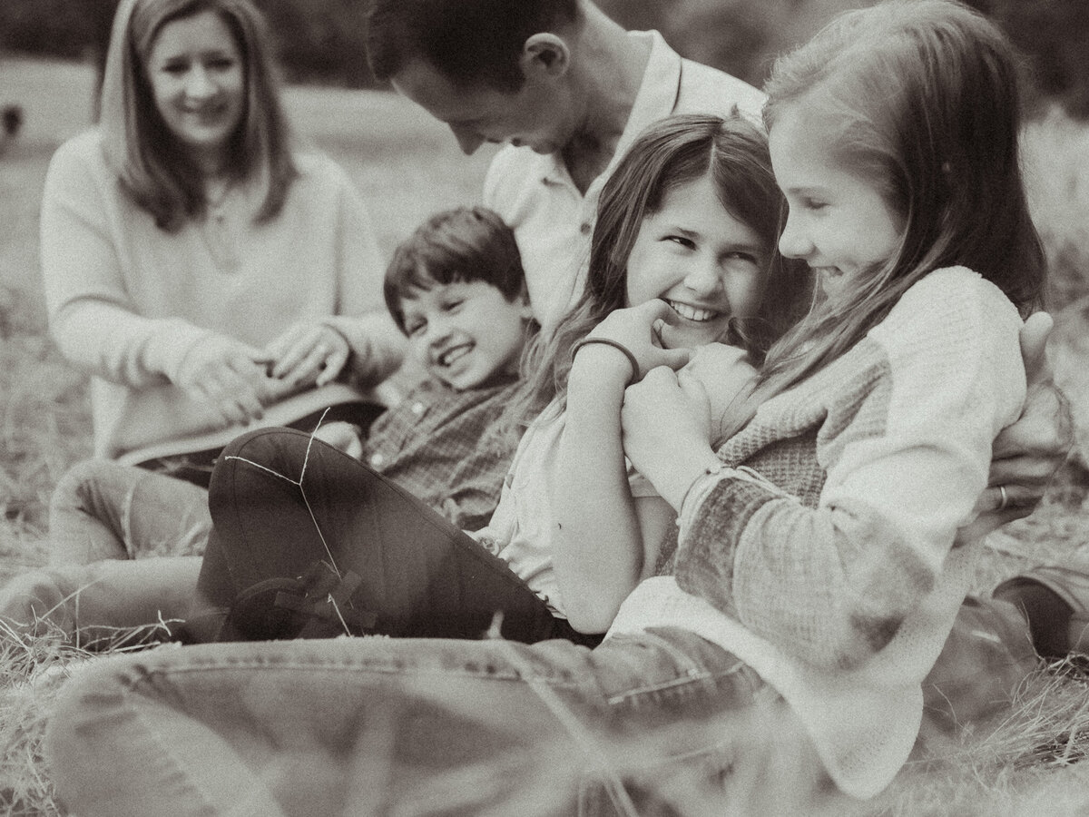 Two sisters giggle together while their family is snuggled up on a blanket in a field at The Vine Photography Venue. 