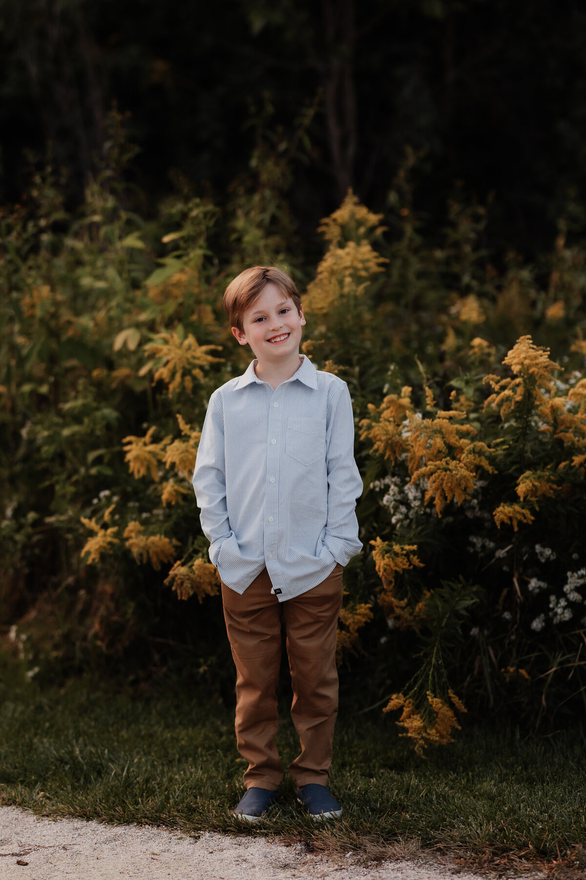A boy smiles with his hands in his pockets in front of yellow flowers.