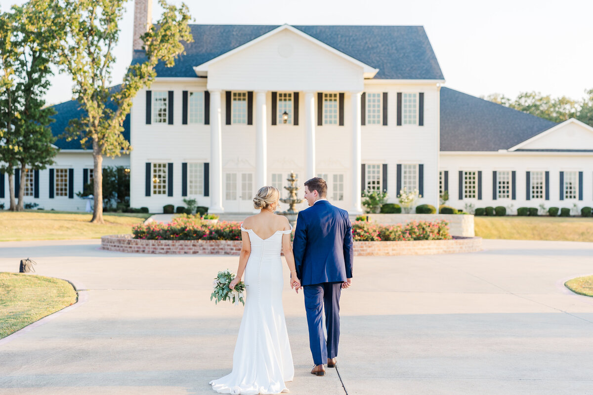 The newlyweds walk hand in hand toward a grand white mansion, capturing a serene and charming moment in Denton, TX.