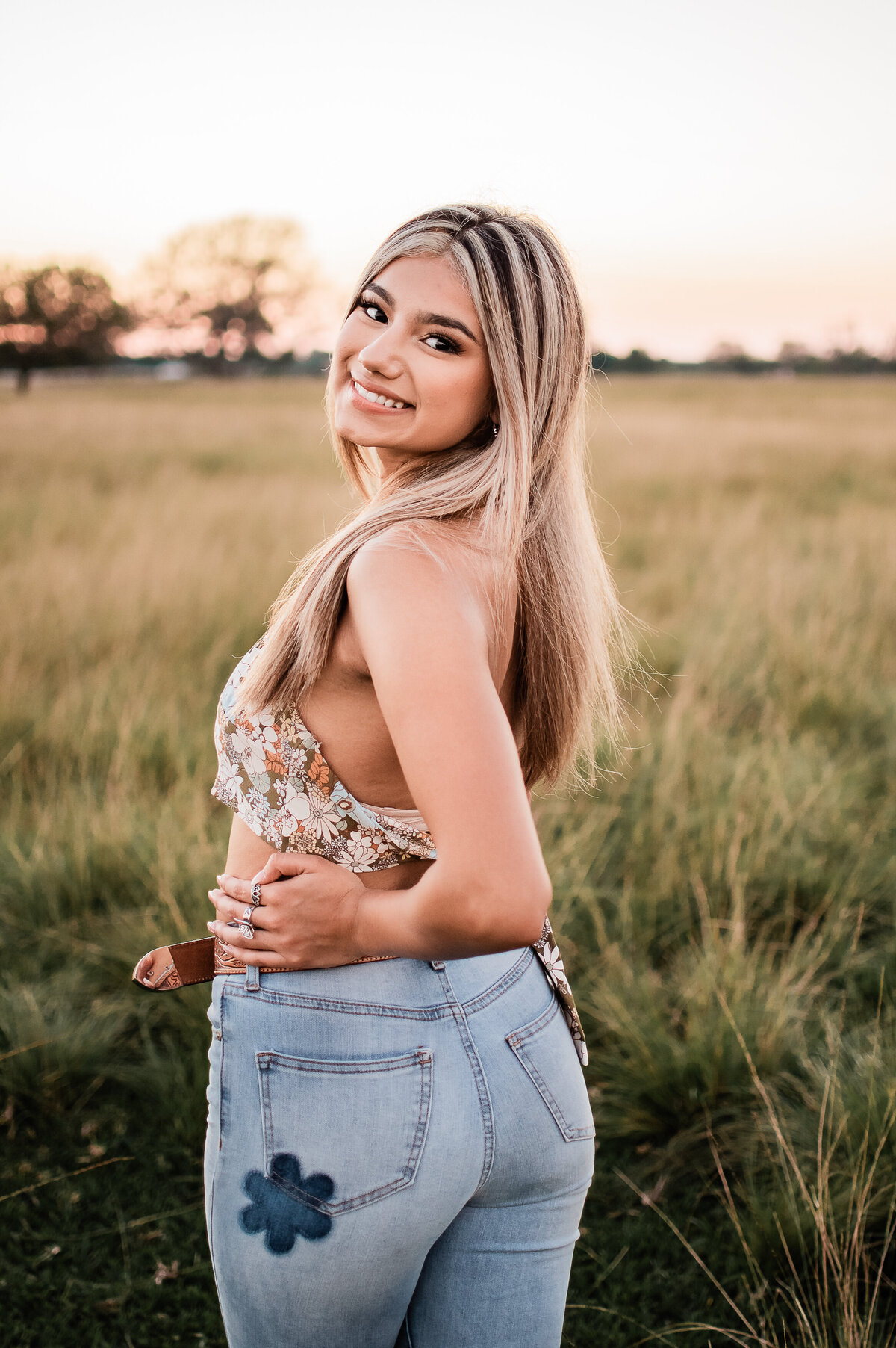 A high school graduate wearing jeans and a floral top looks over her shoulder in a grassy field.