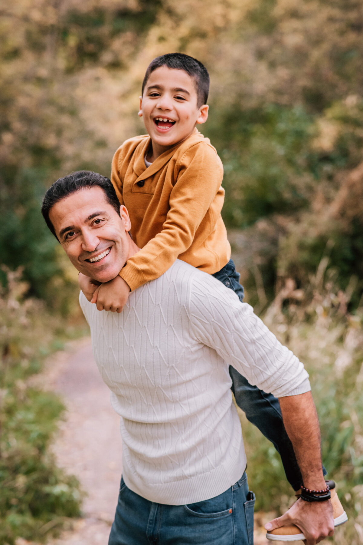 A father and his young son, both smiling brightly, share a joyful moment outdoors. The father is giving the boy a piggyback ride while standing on a nature trail surrounded by greenery.