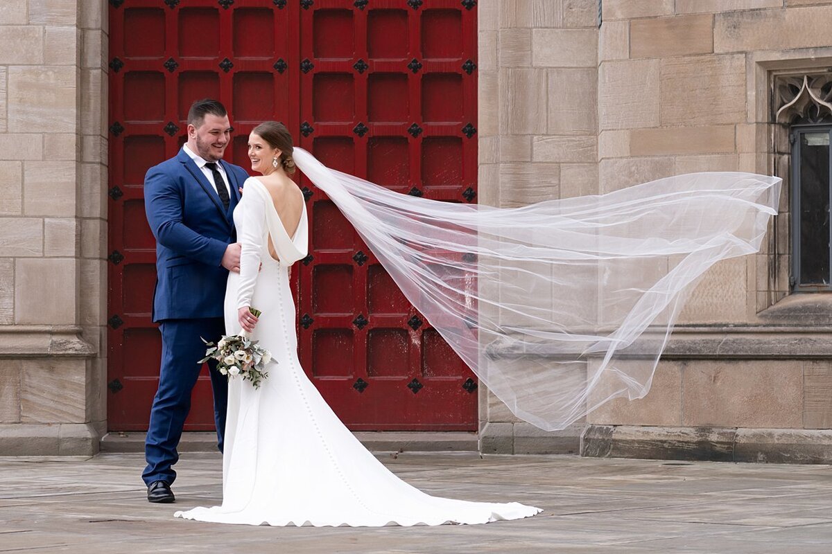 NFL free agent Groom and his Bride with a windswept veil  pose for photos in front of red door at Cathedral of Learning in Pittsburgh, PA