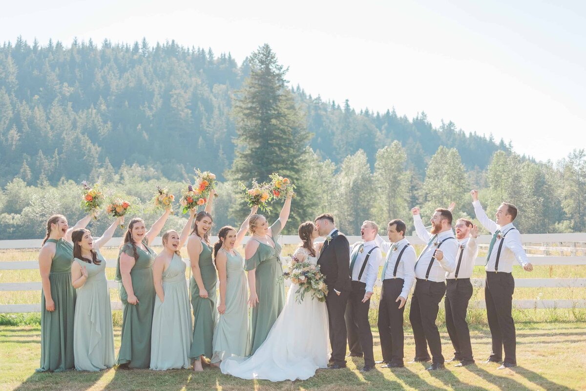 Wedding party of 13 people cheering loudly as the bride and groom kiss in front of them