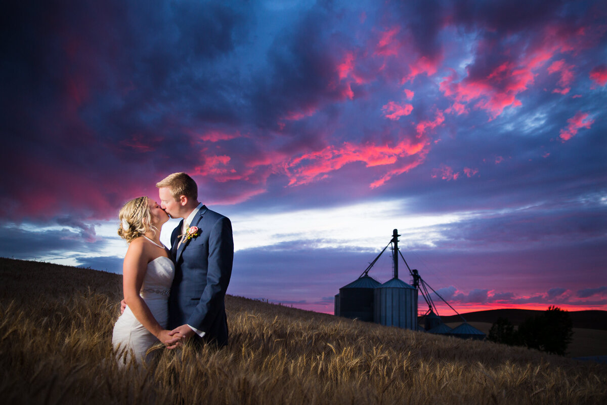 A bride and groom kiss under a colorful sky at sunset during their wedding reception near Pullman, Washington.