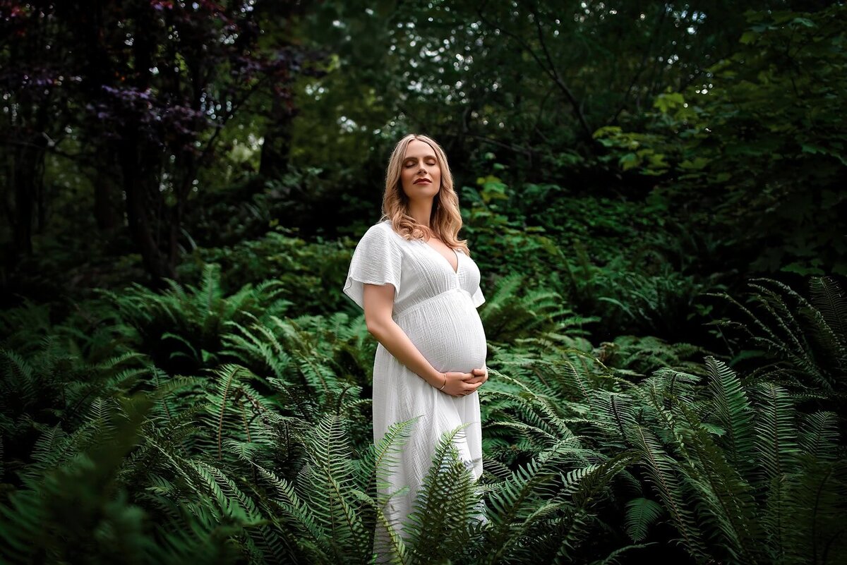 Expectant mom in a white dress surrounded by green ferns during a maternity session at Whytecliff Park in West Vancouver.
