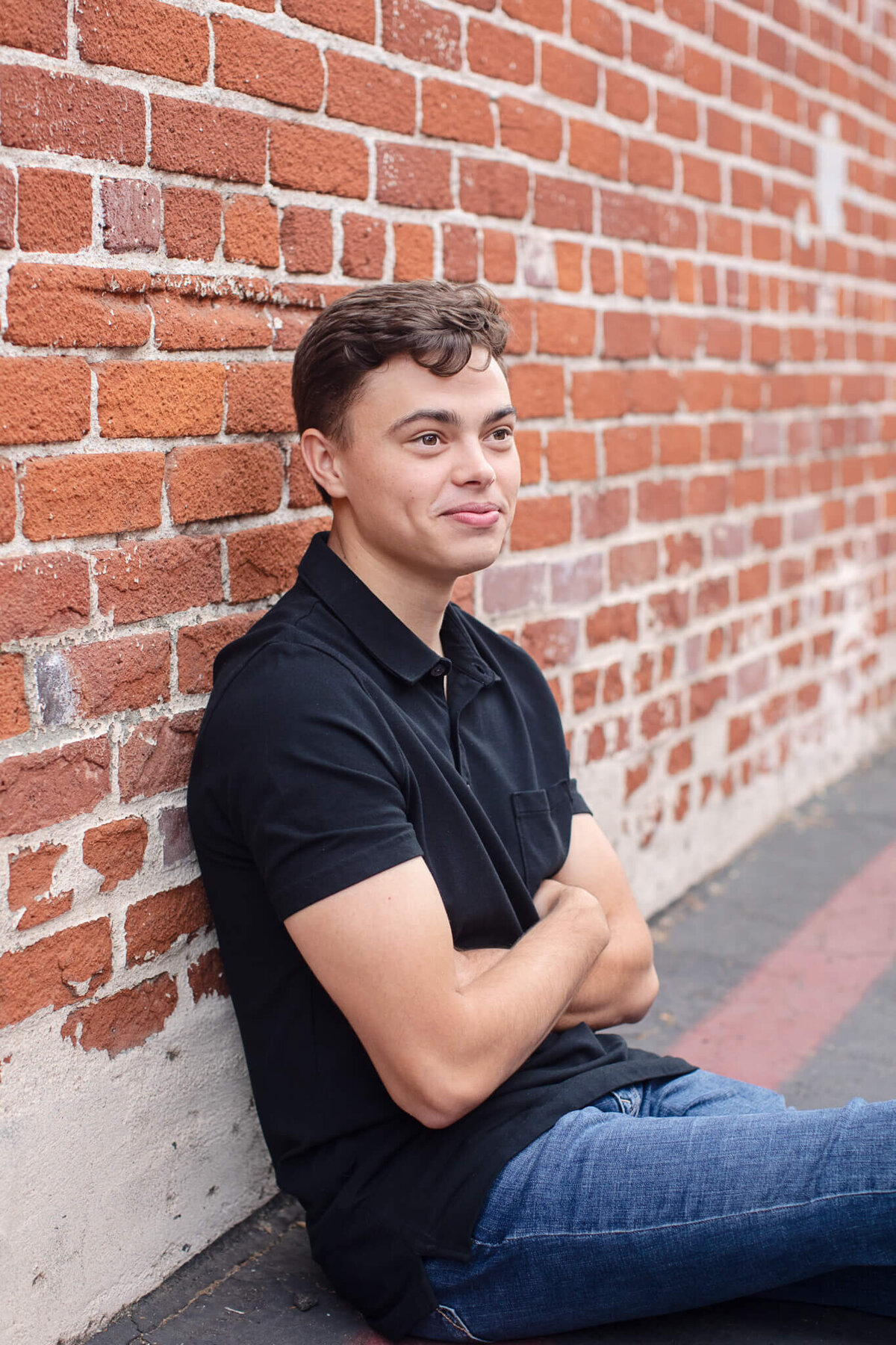 A person wearing a black polo shirt and jeans sits against a red brick wall with their arms crossed. They are looking to the side and have a relaxed expression.