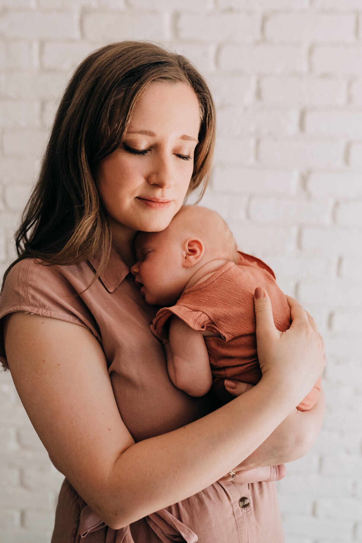 A mother snuggles her newborn daughter against her chest in front of a white brick background during their photoshoot with Boston Newborn Photographer, Allison Wolf