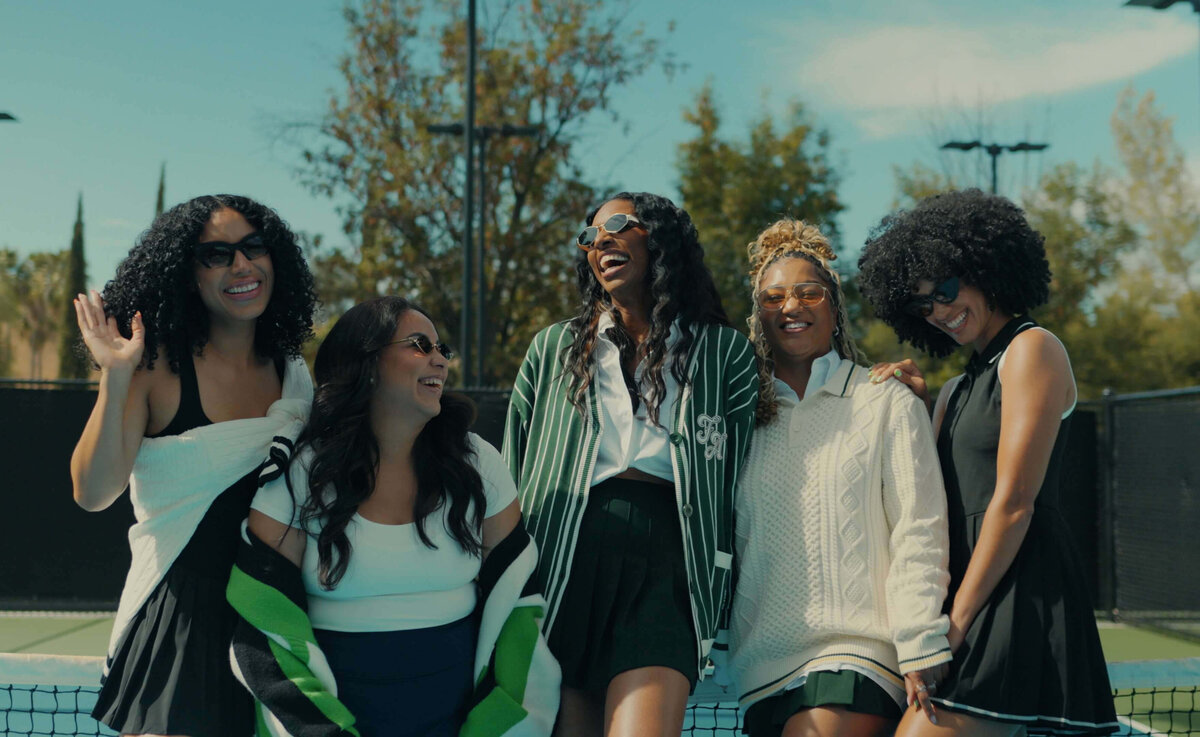 A group of women stood together laughing on a tennis court.