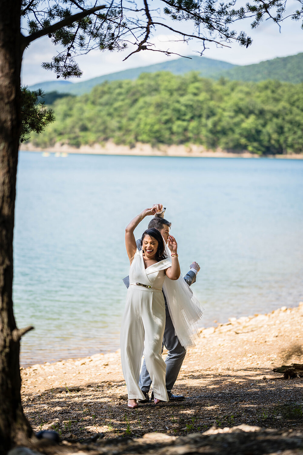 A bride and groom dance along the shores of Carvin’s Cove under a canopy of trees and with the rolling hills of Southwest Virginia behind them. The groom watches closely as they both turn and the bride laughs.