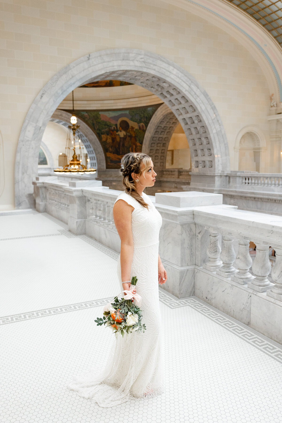 The bride stands with her bouquet in hand, framed by the elegant marble arches of the Utah State Capitol.