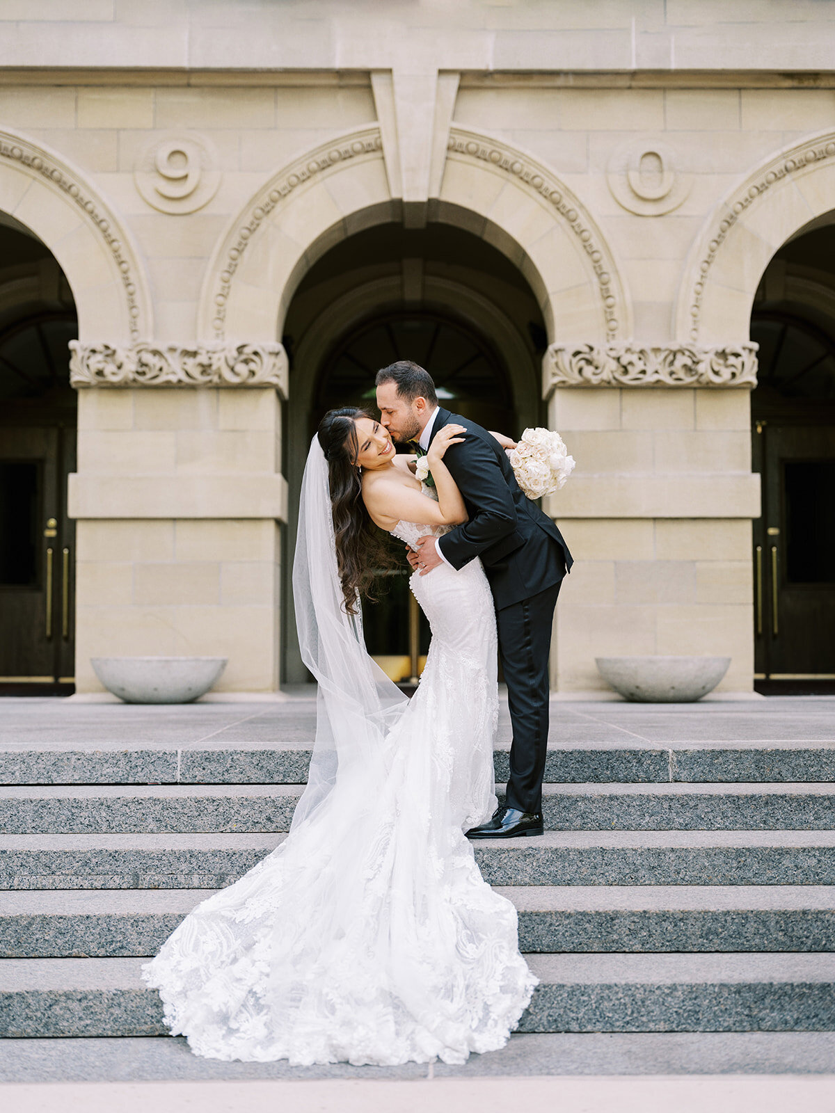 A bride and groom share a kiss on the stone steps in front of an ornate building. The bride holds a bouquet and wears a long, lace dress and veil. The groom is dressed in a dark suit, capturing the elegance of their Fairmont Palliser Wedding Calgary.