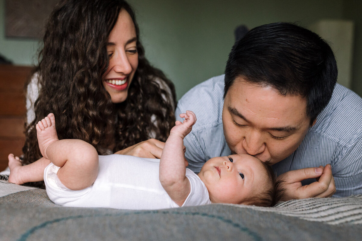 parents laying across bed over newborn in white onesie and dad is kissing baby's head