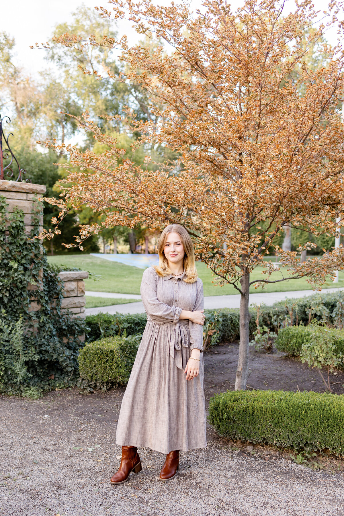 A young woman in a long brown dress stands next to a golden autumn tree in a well-manicured park. She has a soft smile and crosses her arms as she gazes at the camera.