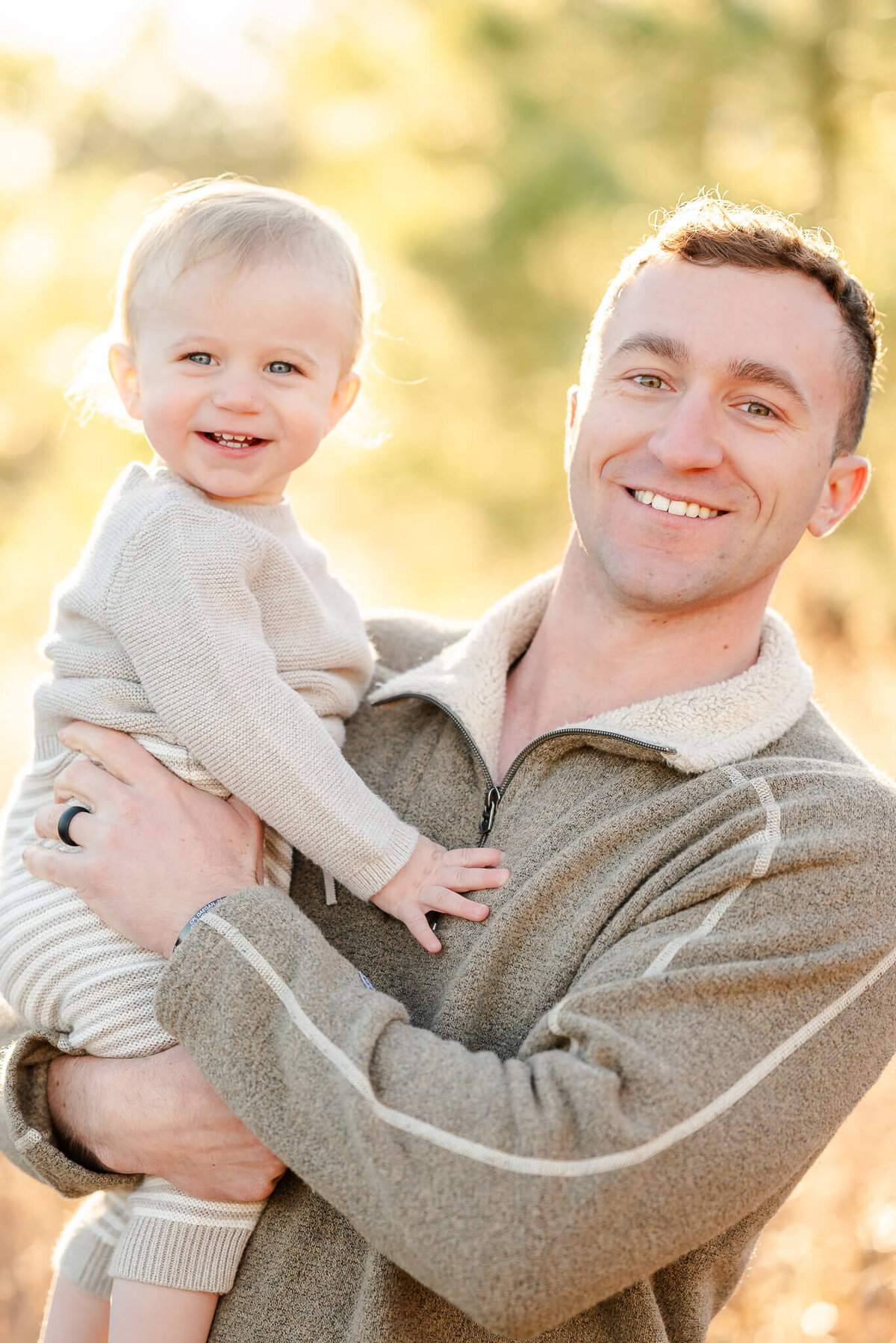 A man wearing a tan sweater holds his toddler son in his arms . The both smile wide while looking at the camera.