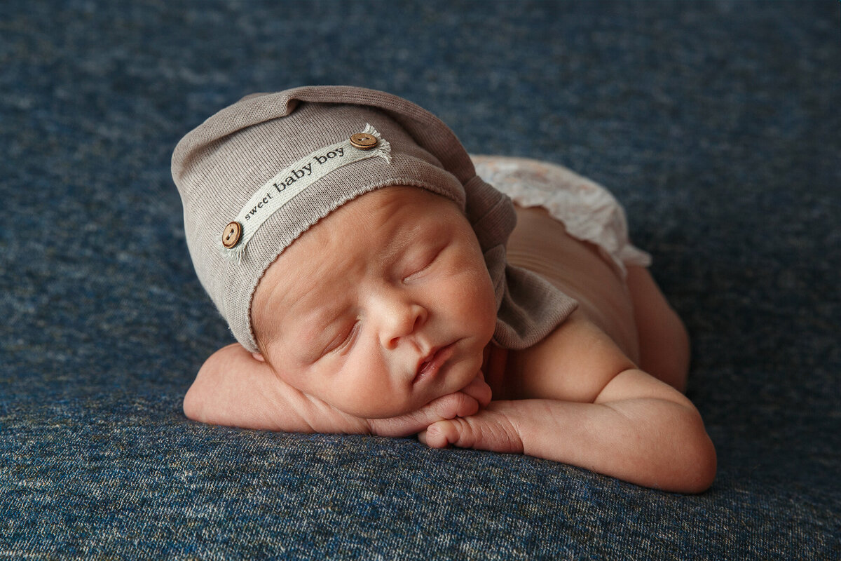 Photograph of a sleeping baby with a little gray night cap sleeping with his chin in his hands on a blue rug